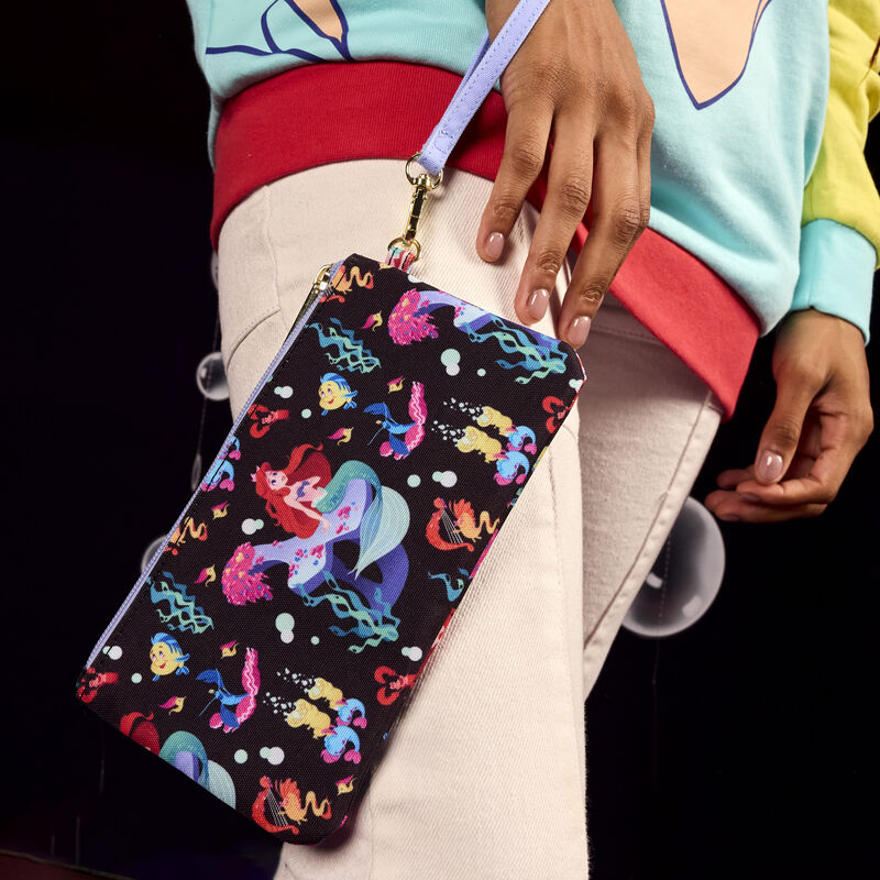 Woman wearing the Loungefly Little Mermaid 35th Anniversary Life is the Bubbles All-Over Print Nylon Zipper Pouch Wristlet around her wrist, featuring an all-over print of Ariel, Flounder, Sebastian, and other aquatic characters and motifs.